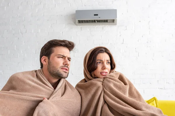 Hombre y mujer congelados calentándose bajo mantas mientras están sentados bajo el aire acondicionado - foto de stock