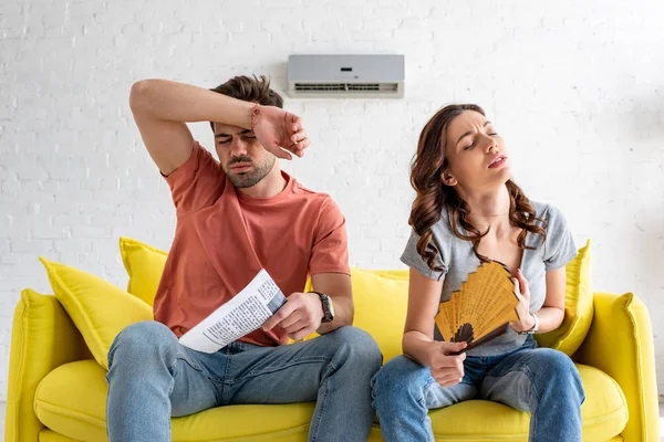 Hübsche Frau mit Handfächer und schöner Mann mit Zeitung, der zu Hause unter Hitze leidet — Stockfoto