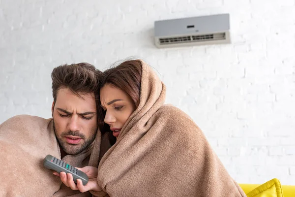 Hübsche Frau hält Fernbedienung in der Hand, während sie sich mit ihrem schönen Freund unter einer Decke wärmt — Stockfoto