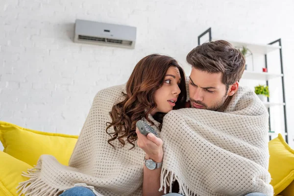 Frozen couple warming under blanket while sitting on sofa under air conditioner at home — Stock Photo