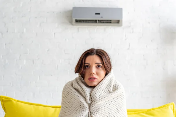 Eingefrorene junge Frau wärmt sich unter Decke, während sie zu Hause auf dem Sofa unter Klimaanlage sitzt — Stockfoto