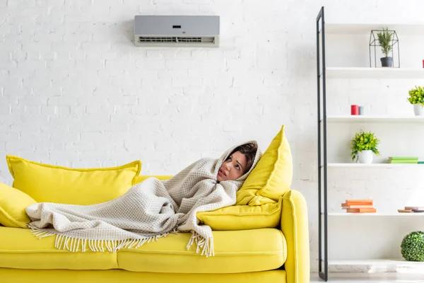 Frozen woman warming with blanket while lying on yellow sofa under air conditioner at home — Stock Photo