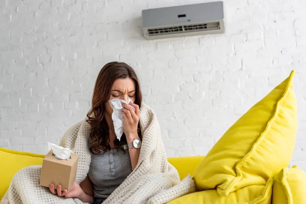 Femme malade avec serviette en papier chauffant sous couverture tout en étant assis sous climatiseur à la maison — Photo de stock