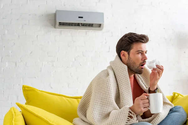 Kranker Mann mit Tasse und Papierserviette wärmt sich unter Decke, während er zu Hause unter Klimaanlage sitzt — Stockfoto
