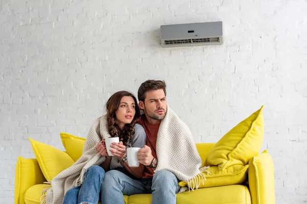 Frozen couple with cups warming under blanket while sitting under air conditioner at home — Stock Photo