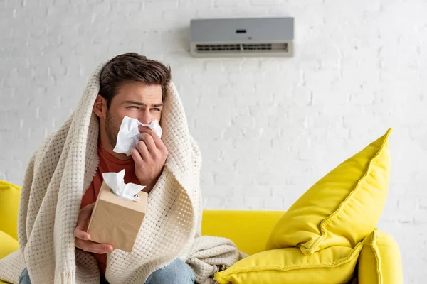 Sick man with paper napkins warming under blanket while sitting under air conditioner at home — Stock Photo