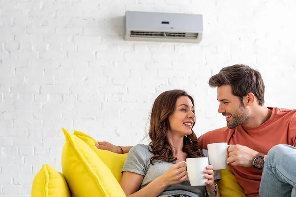Happy couple with cups sitting on sofa under air conditioner at home — Stock Photo