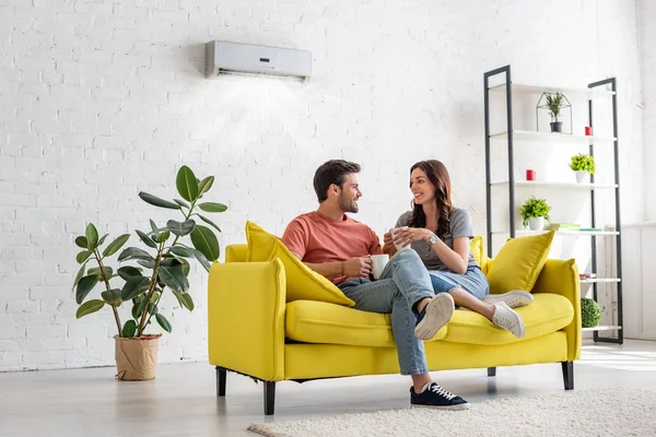 Happy young couple talking and holding cups while sitting on yellow sofa under air conditioner at home — Stock Photo