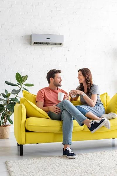 Schöner Mann mit hübscher Freundin, die Tassen hält, während sie zu Hause auf dem Sofa unter der Klimaanlage sitzt — Stockfoto