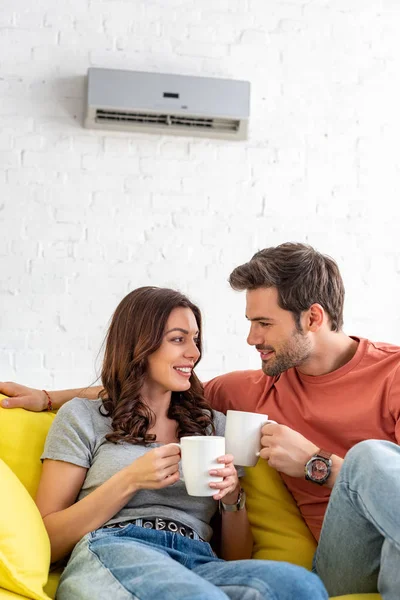 Glückliches lächelndes Paar mit Tassen, während es zu Hause unter einer Klimaanlage sitzt — Stockfoto