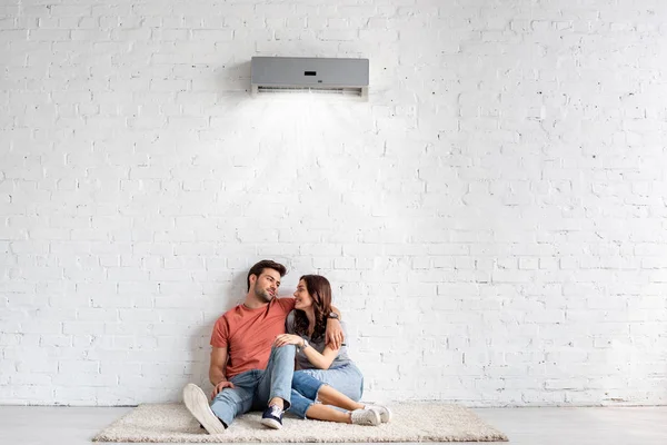 Feliz joven pareja abrazándose mientras está sentado en el suelo cerca de la pared blanca bajo el aire acondicionado - foto de stock