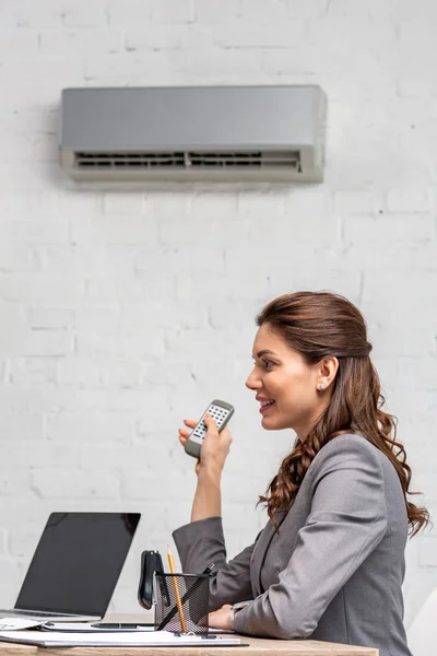 Lächelnde Geschäftsfrau mit Fernbedienung, während sie am Arbeitsplatz unter einer Klimaanlage sitzt — Stockfoto