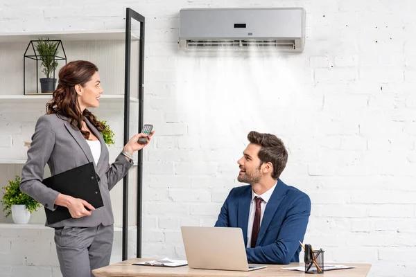 Sonriente mujer de negocios sosteniendo el control remoto mientras está de pie cerca de gerente guapo sentado en el lugar de trabajo bajo el aire acondicionado - foto de stock