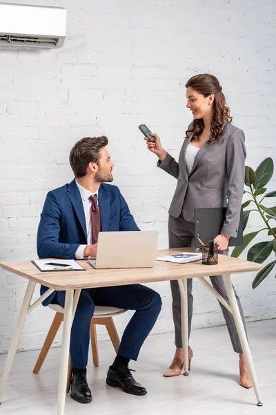 Attrayant femme d'affaires tenant télécommande tout en se tenant près de bel homme en utilisant un ordinateur portable sur le lieu de travail — Photo de stock