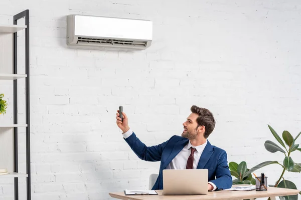 Handsome businessman holding remote control while sitting at workplace under air conditioner — Stock Photo