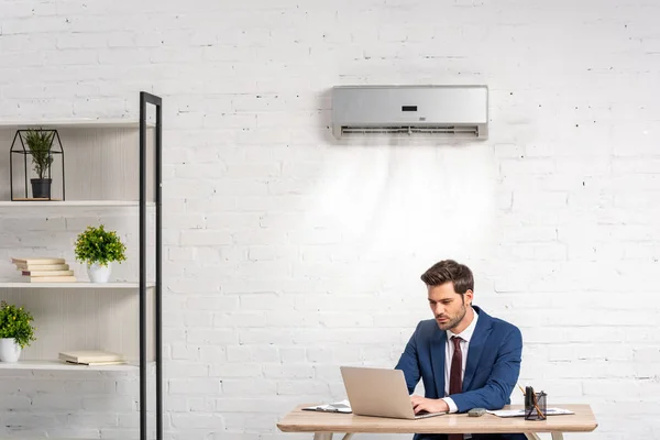 Handsome businessman using laptop while sitting at workplace under air conditioner — Stock Photo