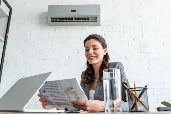 Sonriente mujer de negocios leyendo el periódico mientras está sentado en el lugar de trabajo bajo el aire acondicionado - foto de stock