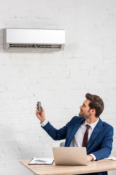 Handsome businessman holding remote control while sitting at workplace under air conditioner — Stock Photo