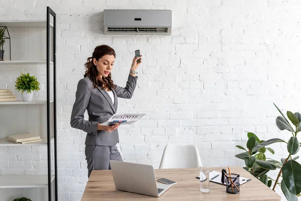 Beautiful businesswoman reading business newspaper while standing under air conditioner with remote control — Stock Photo