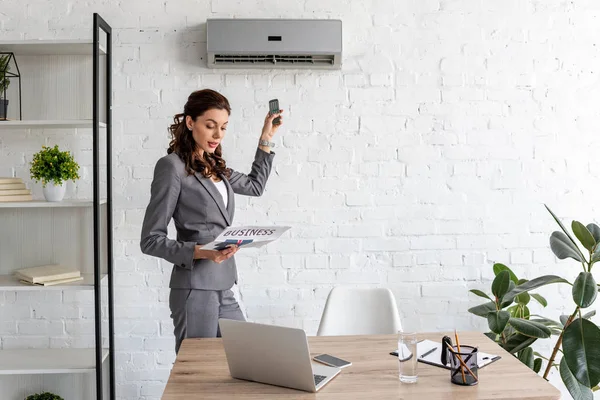 Hermosa mujer de negocios leyendo periódico de negocios, mientras que de pie bajo el aire acondicionado con control remoto - foto de stock