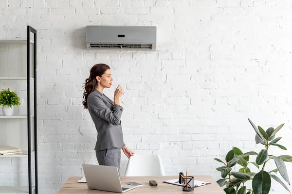 Jolie femme d'affaires eau potable tout en se tenant près du lieu de travail sous climatiseur — Photo de stock