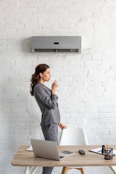 Attractive businesswoman drinking water while standing near workplace under air conditioner — Stock Photo
