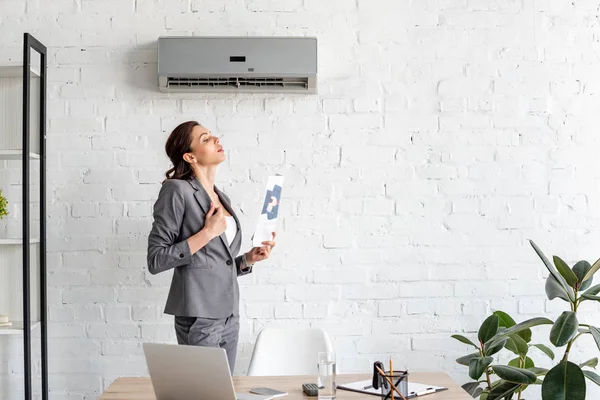 Bonita mujer de negocios agitando el periódico mientras está de pie cerca del lugar de trabajo bajo el aire acondicionado - foto de stock