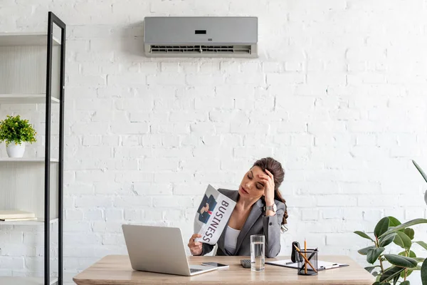 Atractiva mujer de negocios agitando periódico mientras está sentado en el lugar de trabajo bajo el aire acondicionado - foto de stock