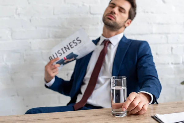 Selective focus of handsome businessman waving business newspaper while sitting at workplace with closed eyes — Stock Photo