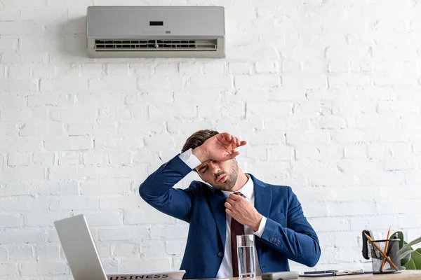 Guapo hombre de negocios que sufre de calor mientras está sentado en el lugar de trabajo bajo el aire acondicionado - foto de stock