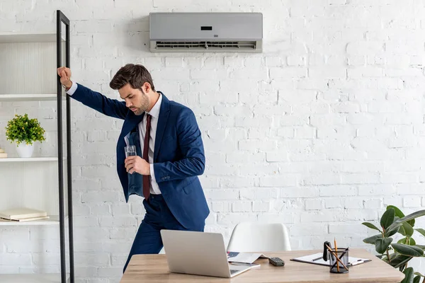 Exhausted businessman holding glass of water while standing near workplace under air conditioner — Stock Photo