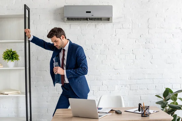 Homme d'affaires épuisé tenant un verre d'eau tout en se tenant près du lieu de travail sous climatiseur — Photo de stock