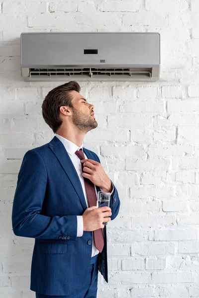 Bel homme d'affaires tenant un verre d'eau tout en se tenant sous le climatiseur — Photo de stock