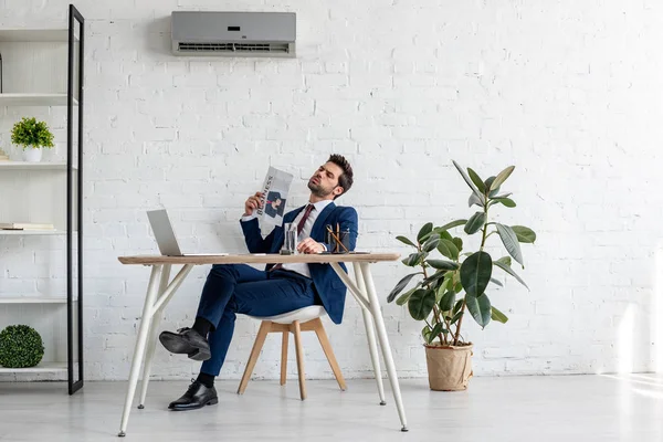 Guapo hombre de negocios agitando periódico de negocios mientras está sentado en el lugar de trabajo y sufriendo de calor en la oficina - foto de stock