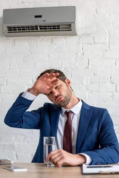 Young businessman suffering from heat while sitting under air conditioner with glass of water — Stock Photo