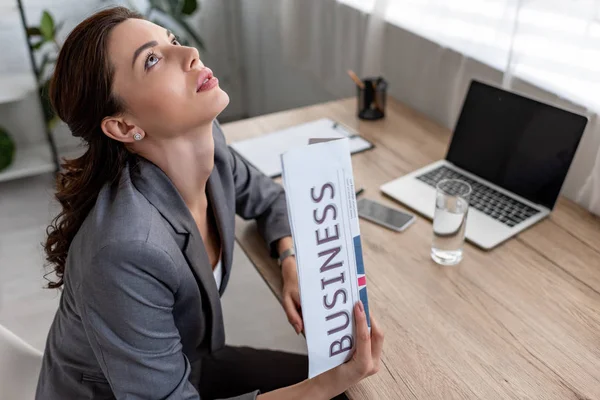 Hübsche Geschäftsfrau winkt mit Wirtschaftszeitung und blickt auf, während sie am Arbeitsplatz im Büro sitzt — Stockfoto