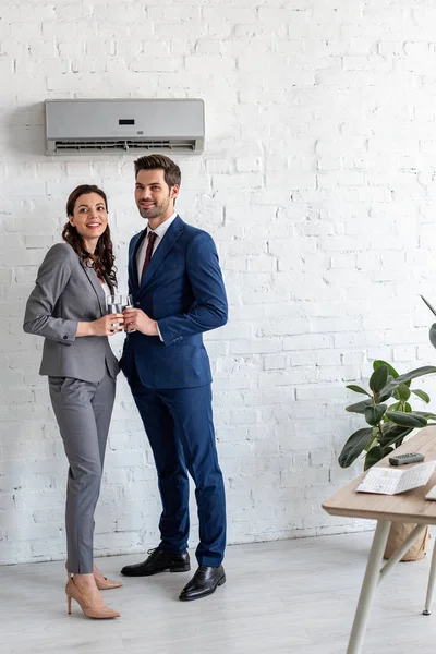 Uomini d'affari sorridenti che tengono bicchieri d'acqua mentre stanno sotto il condizionatore d'aria in ufficio — Foto stock