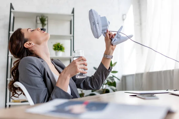 Избирательный фокус привлекательной предпринимательницы, держащей вентилятор и стакан воды, страдающей от жары в офисе — стоковое фото