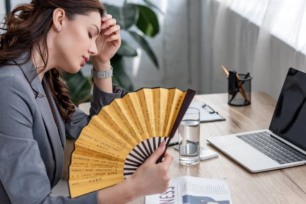 Young businesswoman waving hand fan while sitting at workplace and suffering from heat in office — Stock Photo