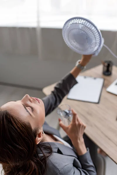 Vista aerea di bella donna d'affari che tiene ventilatore elettrico soffiante e vetro d'acqua mentre soffre di calore in ufficio — Foto stock