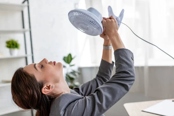 Bonita mujer de negocios con los ojos cerrados sosteniendo ventilador soplado mientras que sufre de calor en la oficina - foto de stock