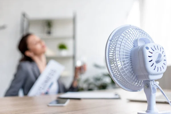 Enfoque selectivo de la mujer de negocios que sufre de calor mientras está sentada cerca de ventilador eléctrico - foto de stock