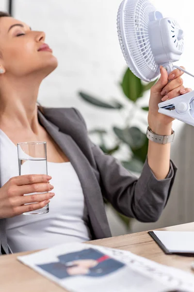 Enfoque selectivo de atractiva empresaria sosteniendo ventilador eléctrico y vaso de agua mientras sufre de calor en la oficina - foto de stock