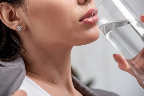Cropped view of young woman suffering from heat and drinking water from glass on grey — Stock Photo