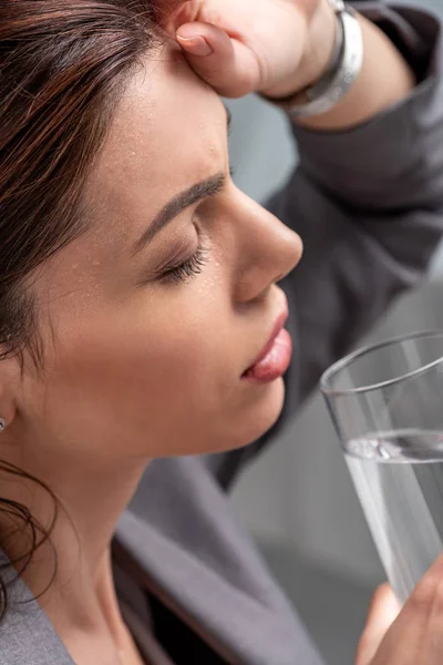Femme épuisée tenant un verre d'eau avec les yeux fermés tout en souffrant de chaleur sur gris — Photo de stock