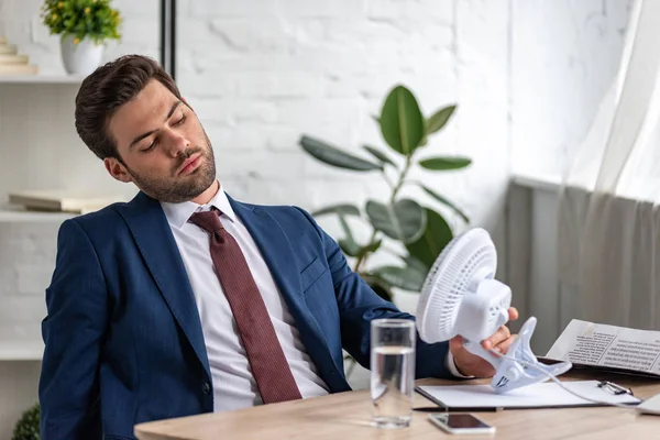 Bonito exausto empresário sofrendo de calor enquanto sentado no local de trabalho na frente de soprar ventilador elétrico — Fotografia de Stock