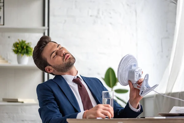 Young businessman holding electric fan while suffering from heat at workplace — Stock Photo