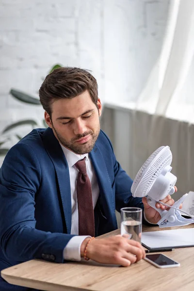 Erschöpfter Geschäftsmann hält am Arbeitsplatz Elektroventilator in der Hand und leidet unter Hitze — Stockfoto