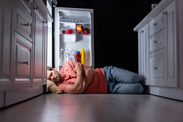 Hombre joven agotado durmiendo en el suelo de la cocina cerca de nevera abierta - foto de stock