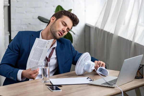 Schöner Geschäftsmann schwenkt Zeitung, während er am Arbeitsplatz vor einem elektrischen Ventilator sitzt und unter Hitze leidet — Stockfoto